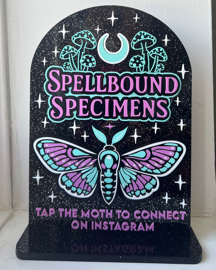 Decorative sign with 'Spellbound Specimens' and a moth design on a black background.