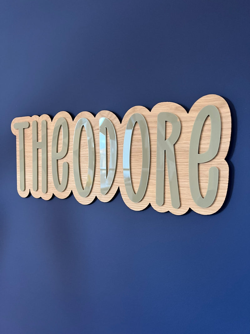 A custom name sign made of oak veneer with the text 'THEODORE' in a cursive font, mounted on a blue wall.