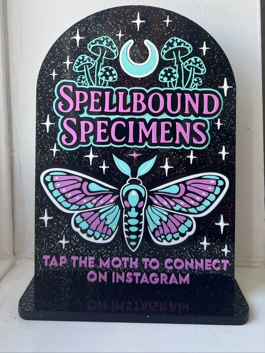 Decorative sign with 'Spellbound Specimens' and a moth design on a black background.