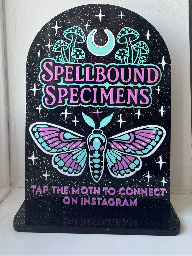 Decorative sign with 'Spellbound Specimens' and a moth design on a black background.