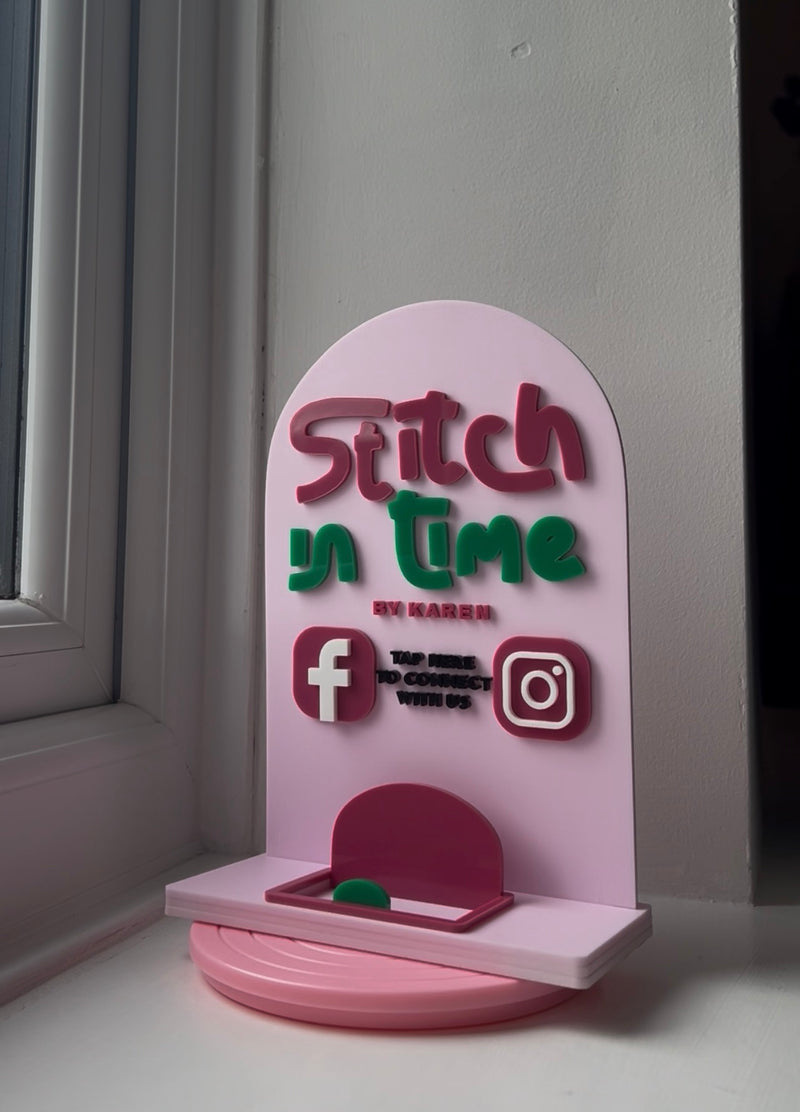 A pink acrylic desk sign with a rounded arch top and base, featuring the text 'Stitch in time' in red and green, along with social media icons for Facebook and Instagram, displayed on a windowsill.